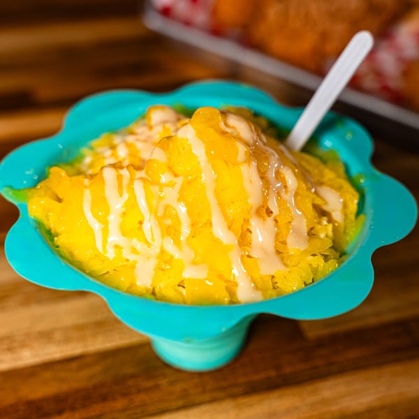 Bright yellow shaved ice dessert with white drizzle in a teal bowl with a white spoon.