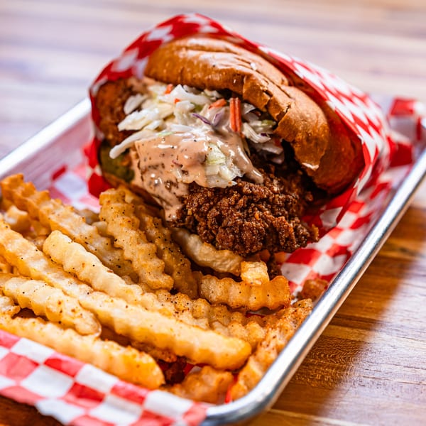 Bird Haus fried chicken sandwich with coleslaw and sauce, served with crinkle-cut fries on a metal tray.