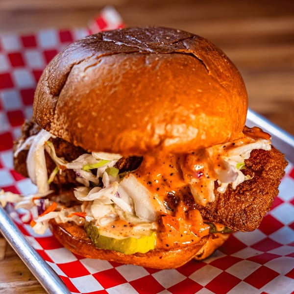 Bird Haus fried chicken sandwich with slaw, pickles, and spicy sauce on a red checkered liner.
