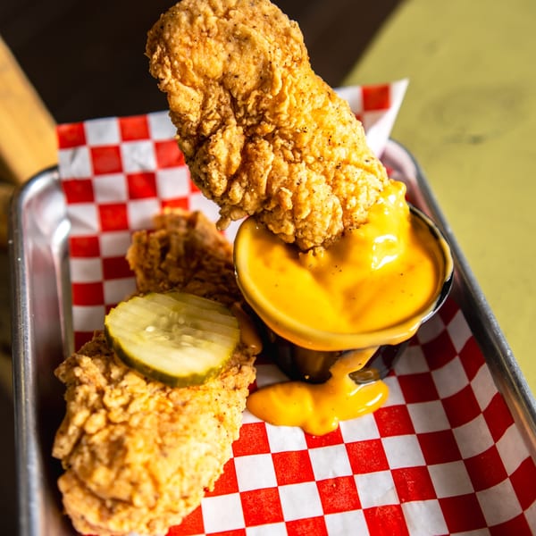Crispy fried chicken tenders with a pickle slice and a side of creamy cheese dipping sauce.