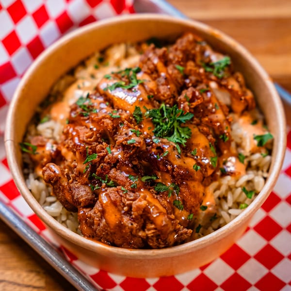 Bird Haus fried chicken and rice bowl, drizzled with sauce and garnished with parsley, on a red checkered liner.